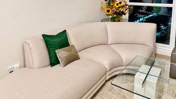 Un sofá color beige con un cojín verde y dorado, una mesa de centro de cristal y un jarrón con flores secas.