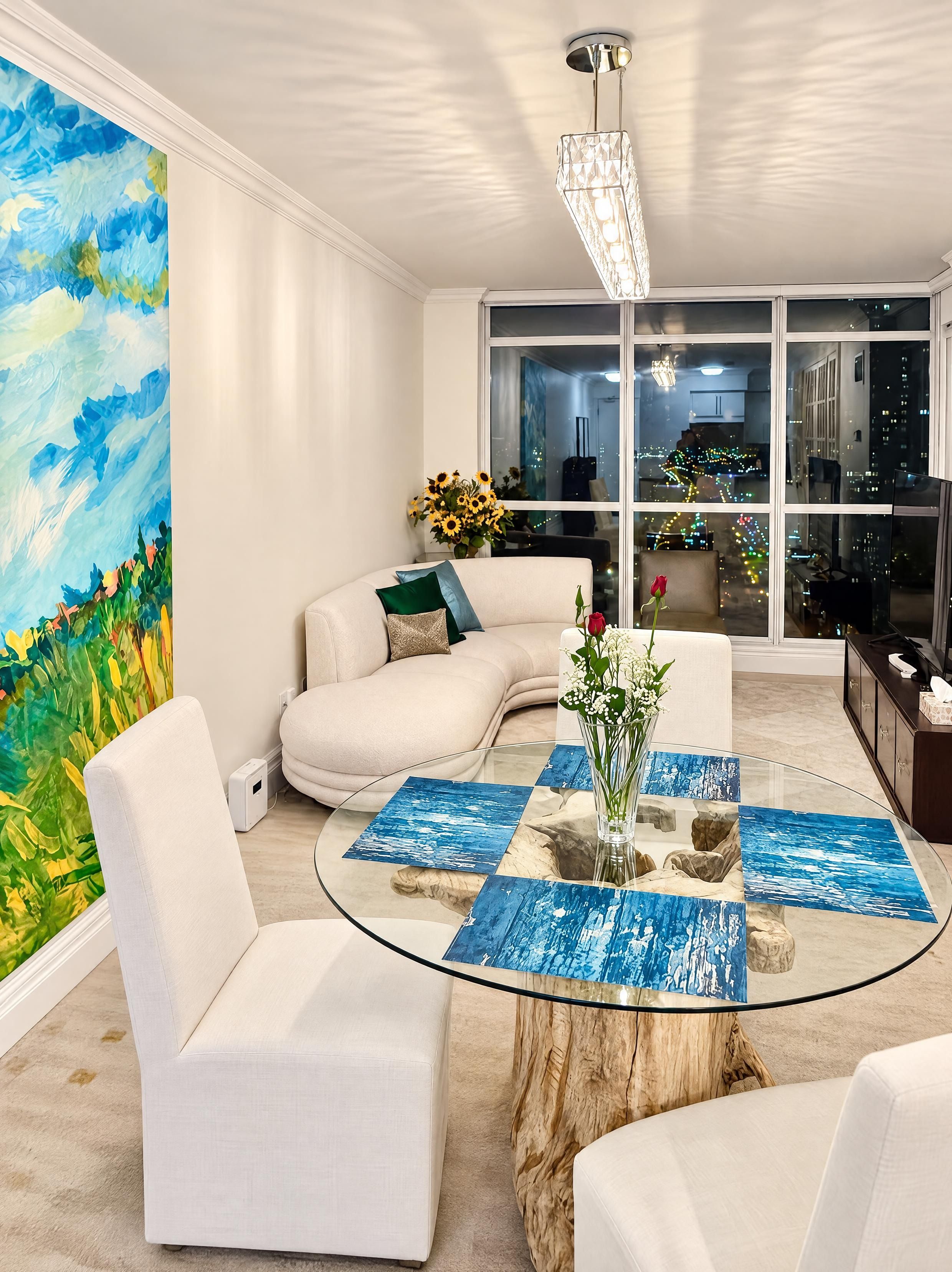 Una sala de estar moderna con una mesa de centro de vidrio, sofás blancos, un cuadro abstracto grande y vistas de la ciudad a través de amplias ventanas.