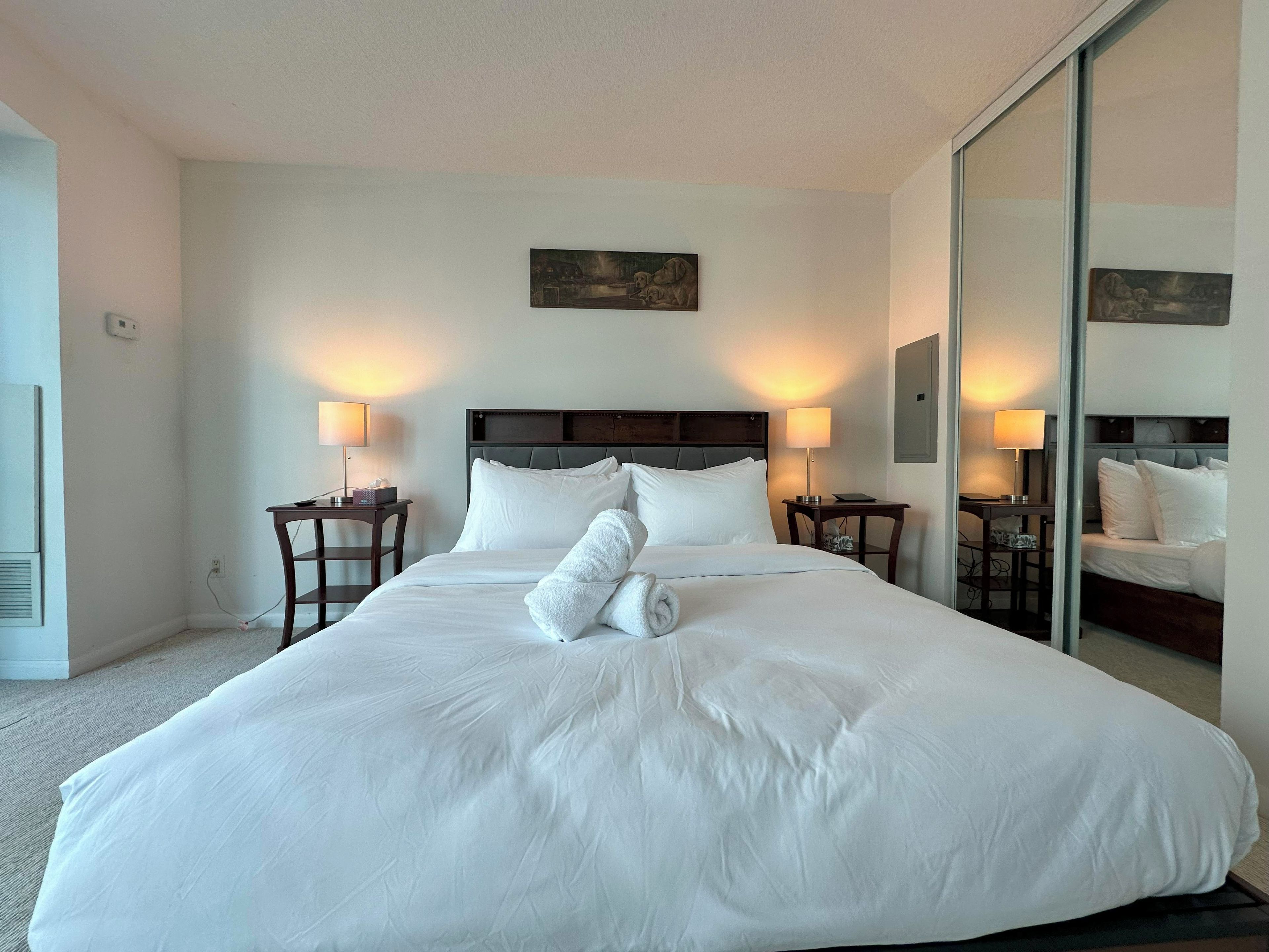 Habitación de hotel con una cama grande, dos mesitas de noche con lámparas, un cuadro en la pared y un espejo que refleja la cama.