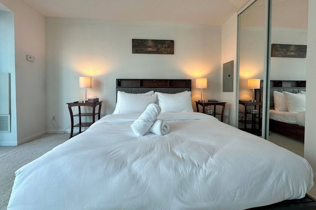 A hotel room with a large bed, two bedside tables with lamps, a painting on the wall, and a mirror reflecting the bed.
