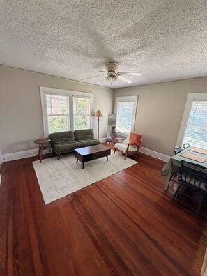 A spacious living room with hardwood flooring, a green sofa, a coffee table, a dining table with chairs, and a ceiling fan. - Historic Riverside Brick Deco | Charming Jax Getaway (Jacksonville)