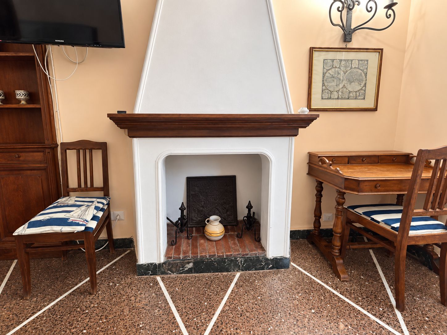 A room with a fireplace, two wooden chairs with striped cushions, a wooden table, a television on the wall, and a framed map.