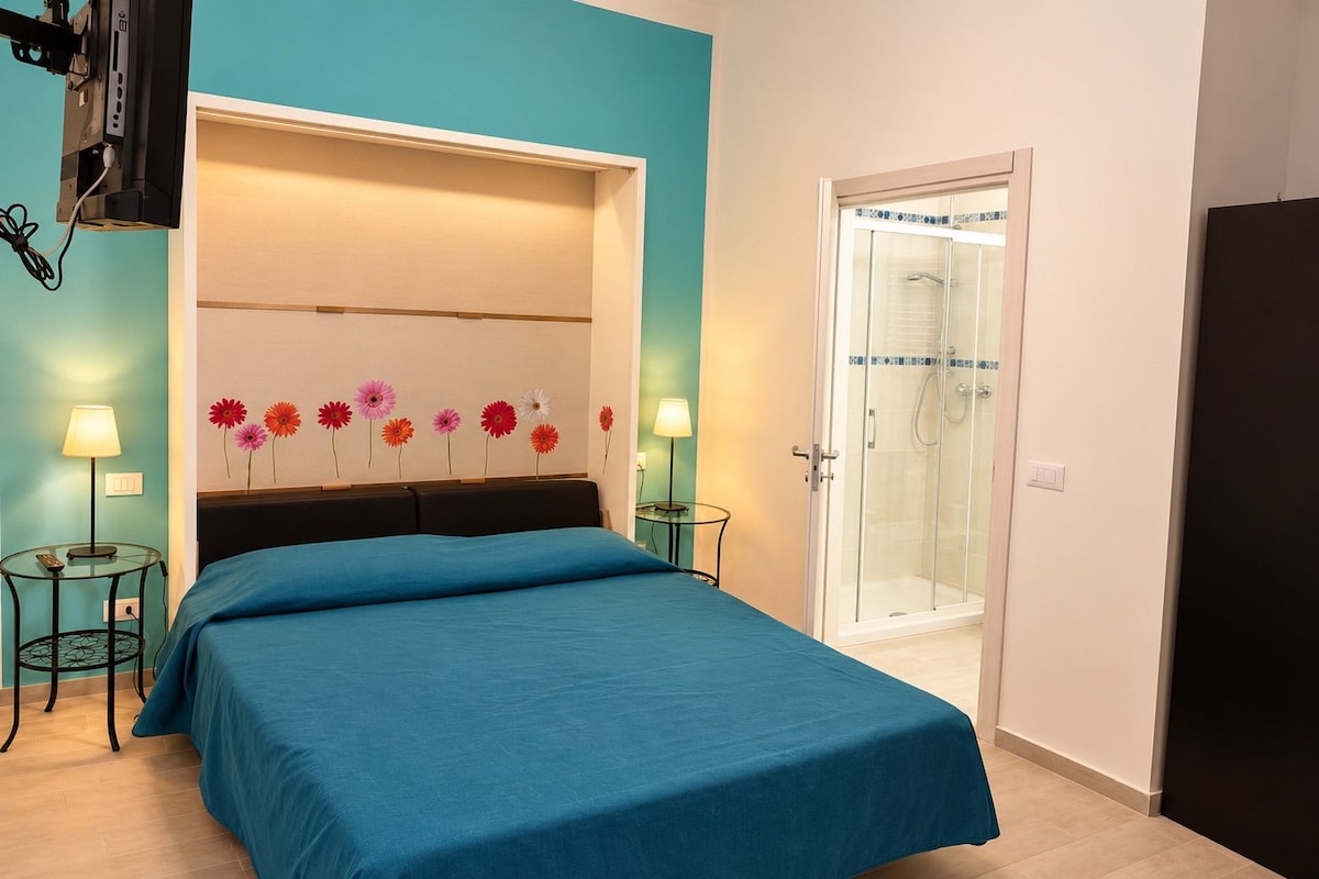 Een slaapkamer met een bed, nachtkastje, lamp, douchegedeelte en een muur met bloemendecoraties.