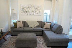 Una sala de estar con un sofá seccional gris, un reposapiés, una alfombra estampada y un cuadro colgado en la pared con flores blancas.