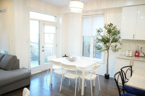 Une cuisine moderne avec des meubles blancs, une table à manger avec des chaises, une plante en pot et un canapé gris.