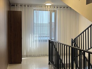 Un pasillo con puerta de madera, cortinas translúcidas, una escalera con barandilla negra y vista a un edificio.