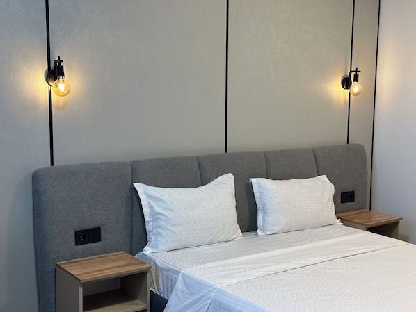 A modern bedroom with a grey upholstered headboard, two bedside tables, and a bed with white bedding.