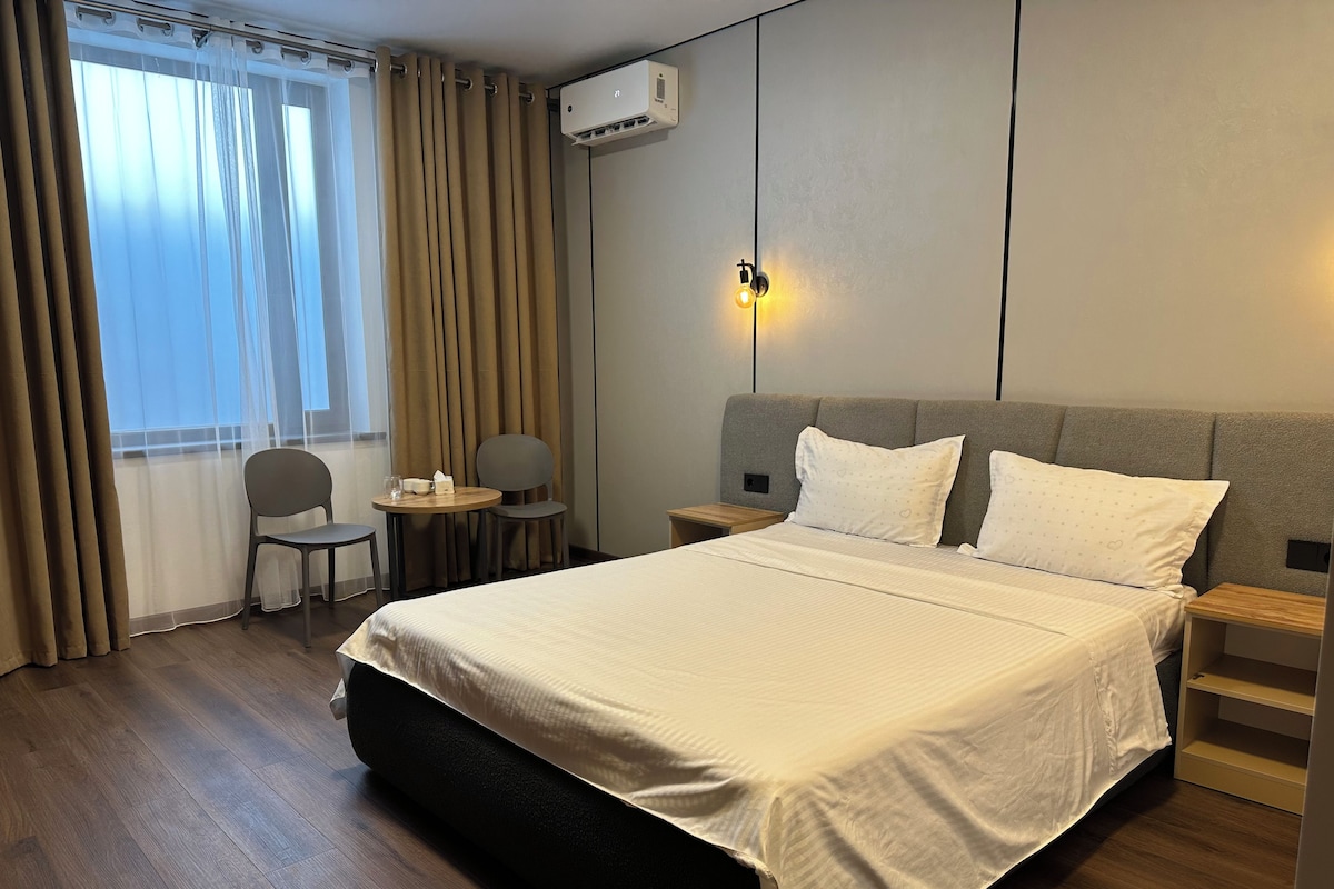 Hotellihuone, jossa on suuri sänky, yöpöytä, tuoli, pieni pöytä lampun kanssa ja ikkuna, jossa on verhot.