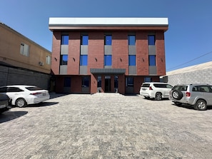 A modern building with a brick facade, multiple windows, and a flat roof. There are several cars parked in front.