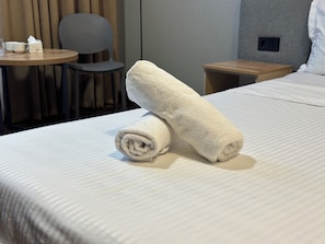 Siististi pedattu sänky, jossa on kaksi rullalle taitettua pyyhettä ja pieni pöytä, jonka päällä on kuppi ja paperinenäliöpakkaus.