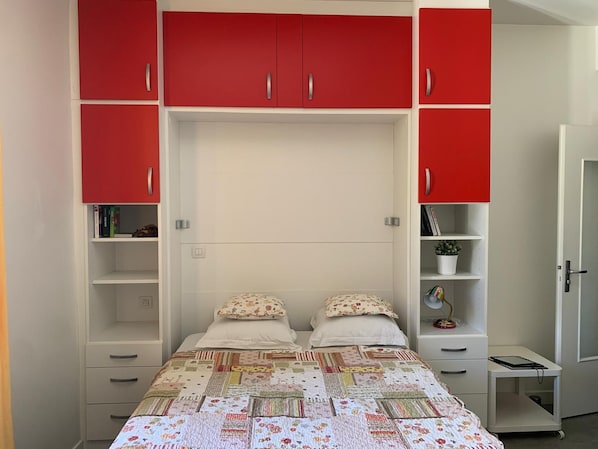 Un dormitorio con una cama, dos mesitas de noche, un armario con puertas rojas y un escritorio pequeño con una silla.