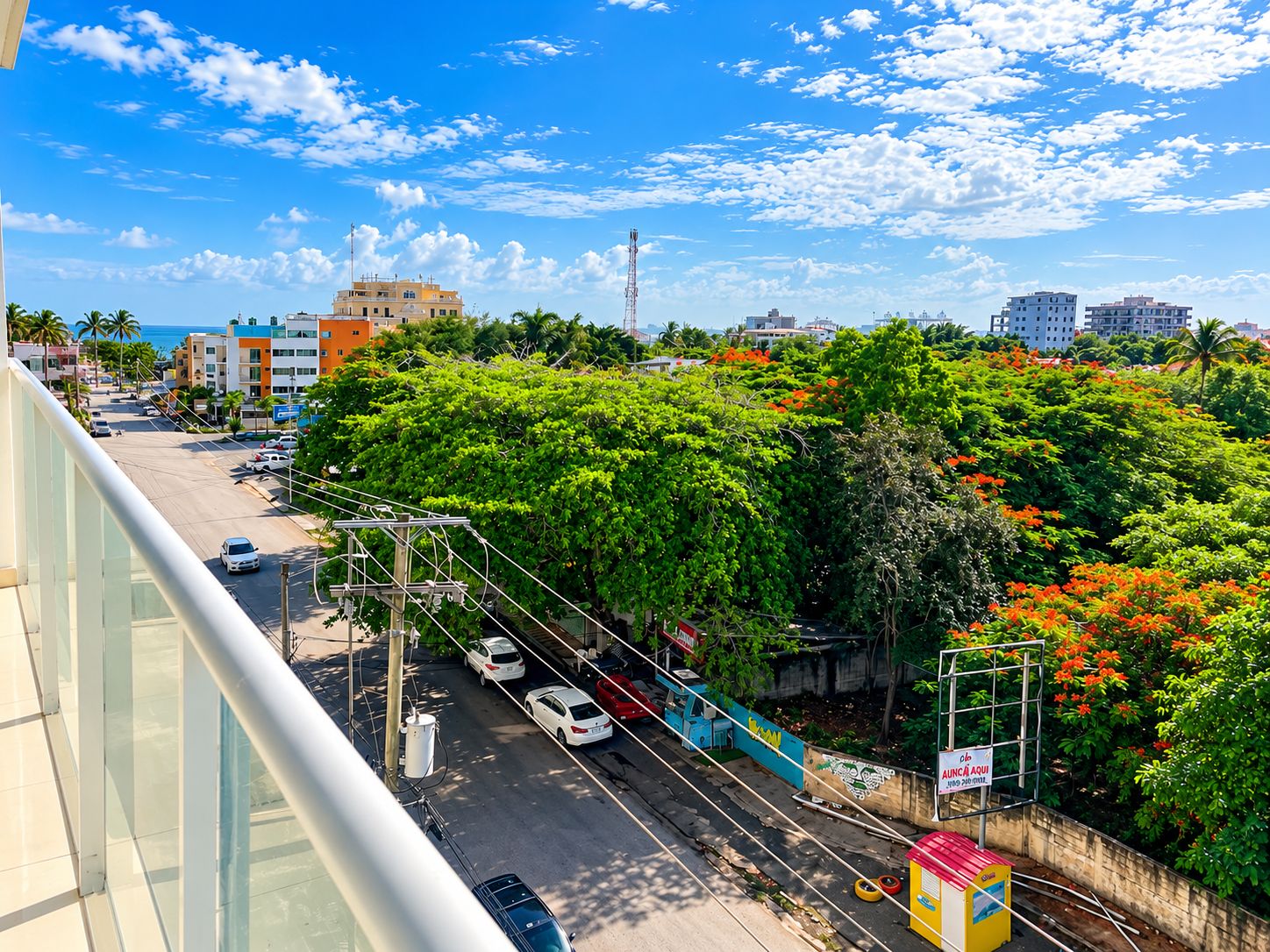 A cityscape with a clear sky, a street with parked cars, and a dense green area with trees.