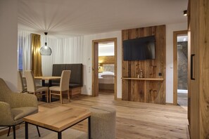 Ein modernes Hotelzimmer mit Holzboden, einer Essecke mit Tisch und Stühlen, einem Sofa, einem an einer Holzplatte montierten Fernseher und einem Bett mit Kopfteil.