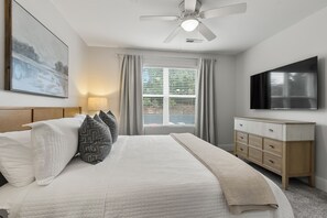 Une chambre à coucher comprenant un lit, un ventilateur de plafond, une fenêtre avec des rideaux, une table de chevet et un téléviseur à écran plat fixé au mur.