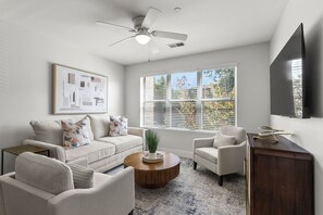 Un salon doté d’un ventilateur de plafond, d’une grande fenêtre avec stores, d’un téléviseur à écran plat fixé au mur et d’un tableau abstrait encadré.