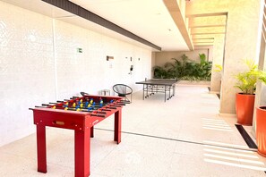 A foosball table and a ping pong table in a hallway with tiled walls and potted plants. - FR Bueno de Alto Padrão Lazer Premium (Goiânia)
