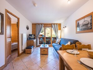 A cozy living room with a sofa, a dining table with chairs, a television, and a view of a balcony with outdoor furniture. - Bright apartment near slopes - 6 guests (Les Menuires)