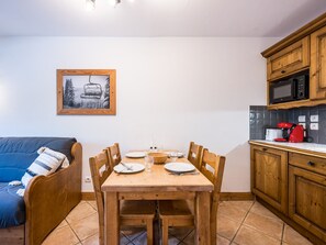Zona de menjador amb taula de fusta per a dues persones, sofà blau i cuina amb armaris de fusta i microones.
