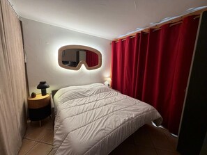 A bedroom with a bed, a nightstand, a lamp, a mirror, and red curtains. - Bright apartment near the slopes (Les Menuires)