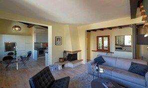 Una sala de estar moderna con un sofá, una mesa de centro de vidrio y una chimenea. Hay una zona de comedor con una mesa y sillas de vidrio, y una cocina al fondo.