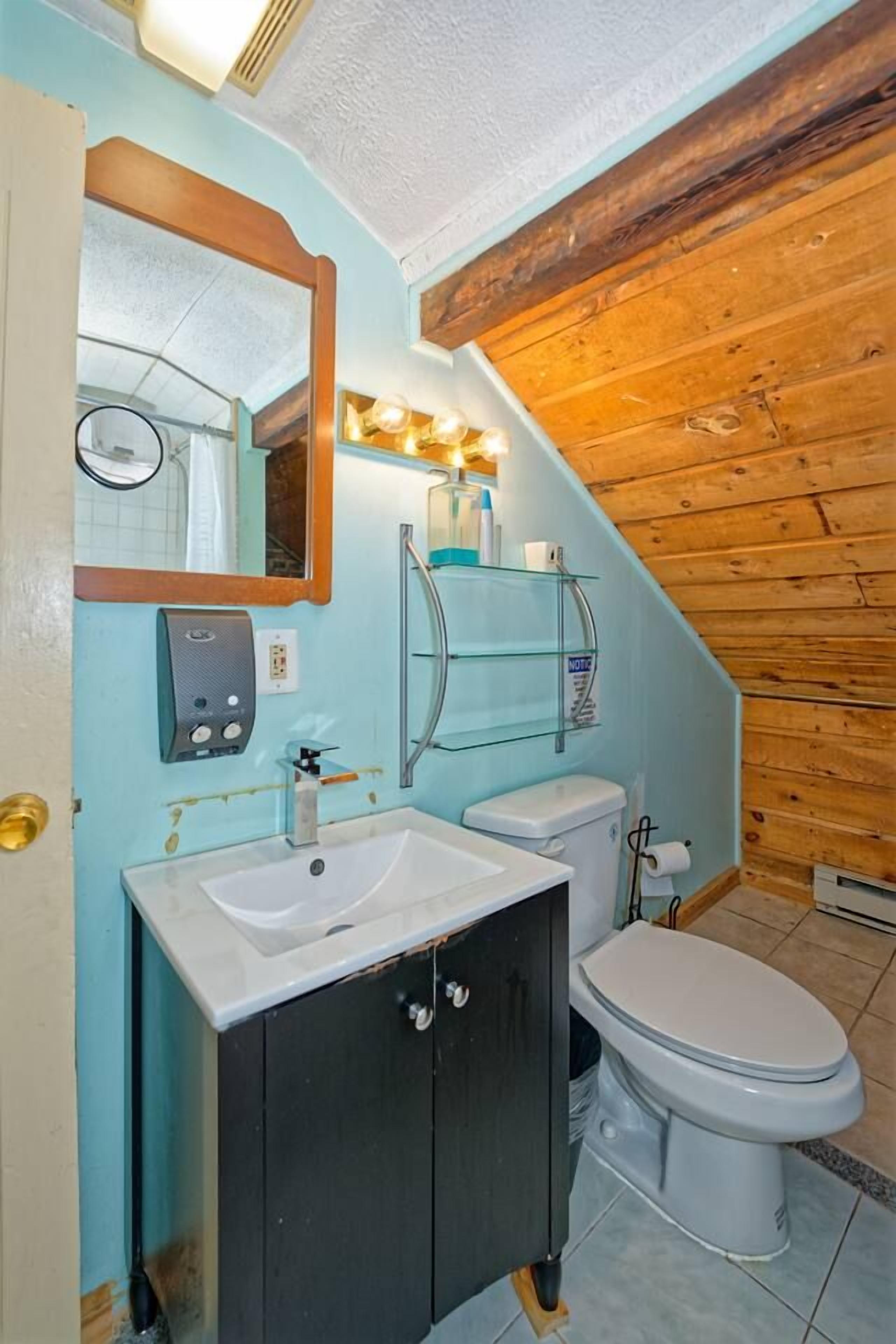 A bathroom with a white sink, dark cabinets, a toilet, a glass shelf with toiletries, and a mirror with lights.