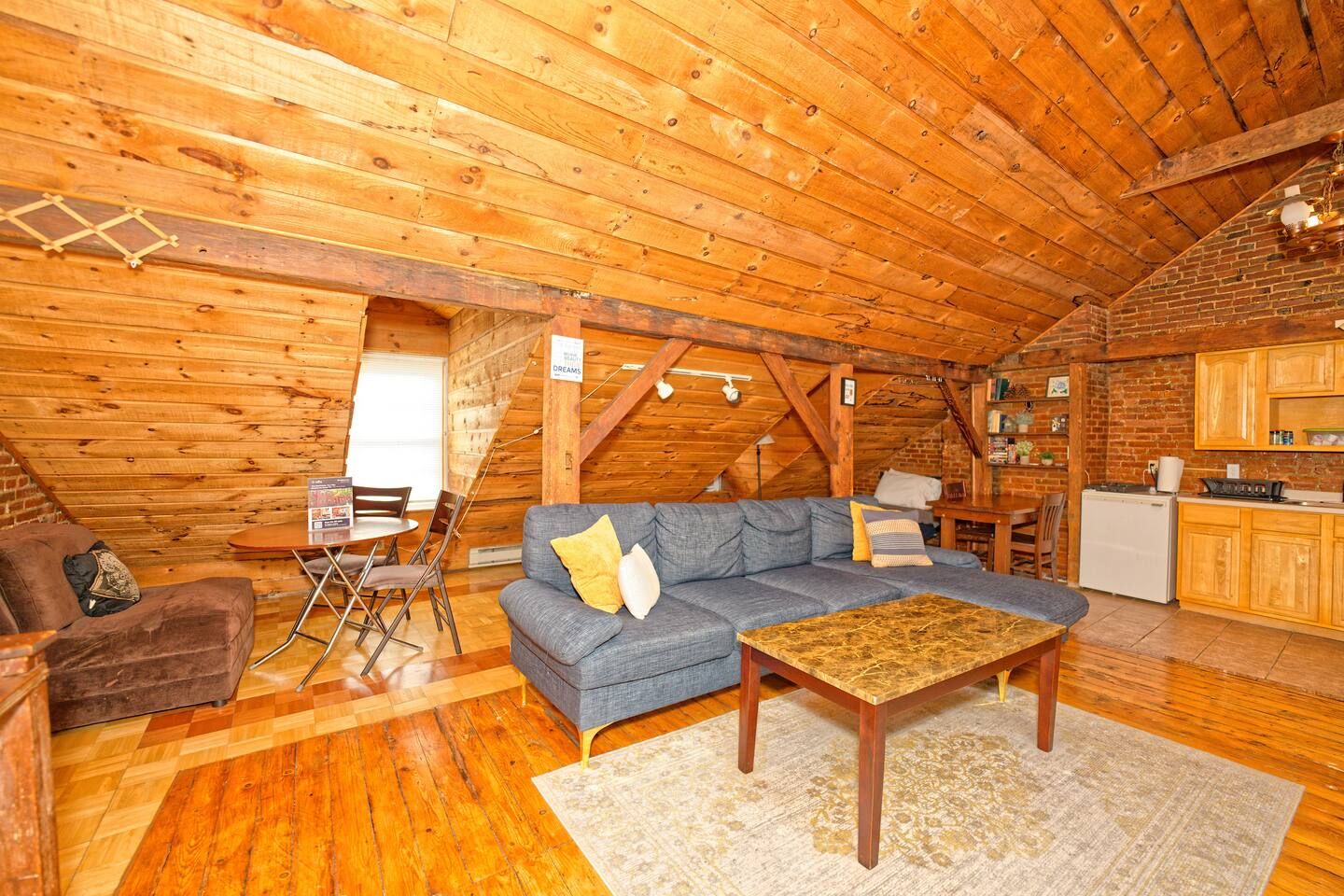 A cozy attic room with a wooden ceiling, a sofa, a coffee table, a dining table, and a kitchen area.
