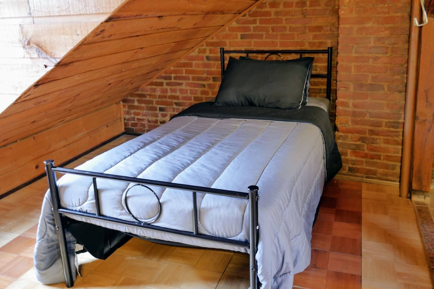 A single bed with a metal frame and a grey mattress in a room with a sloped ceiling and exposed brick walls.