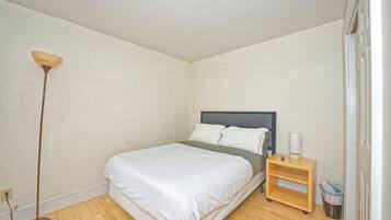 Una cama individual con cabecera gris, una mesita de noche con lámpara y un cubo de basura.