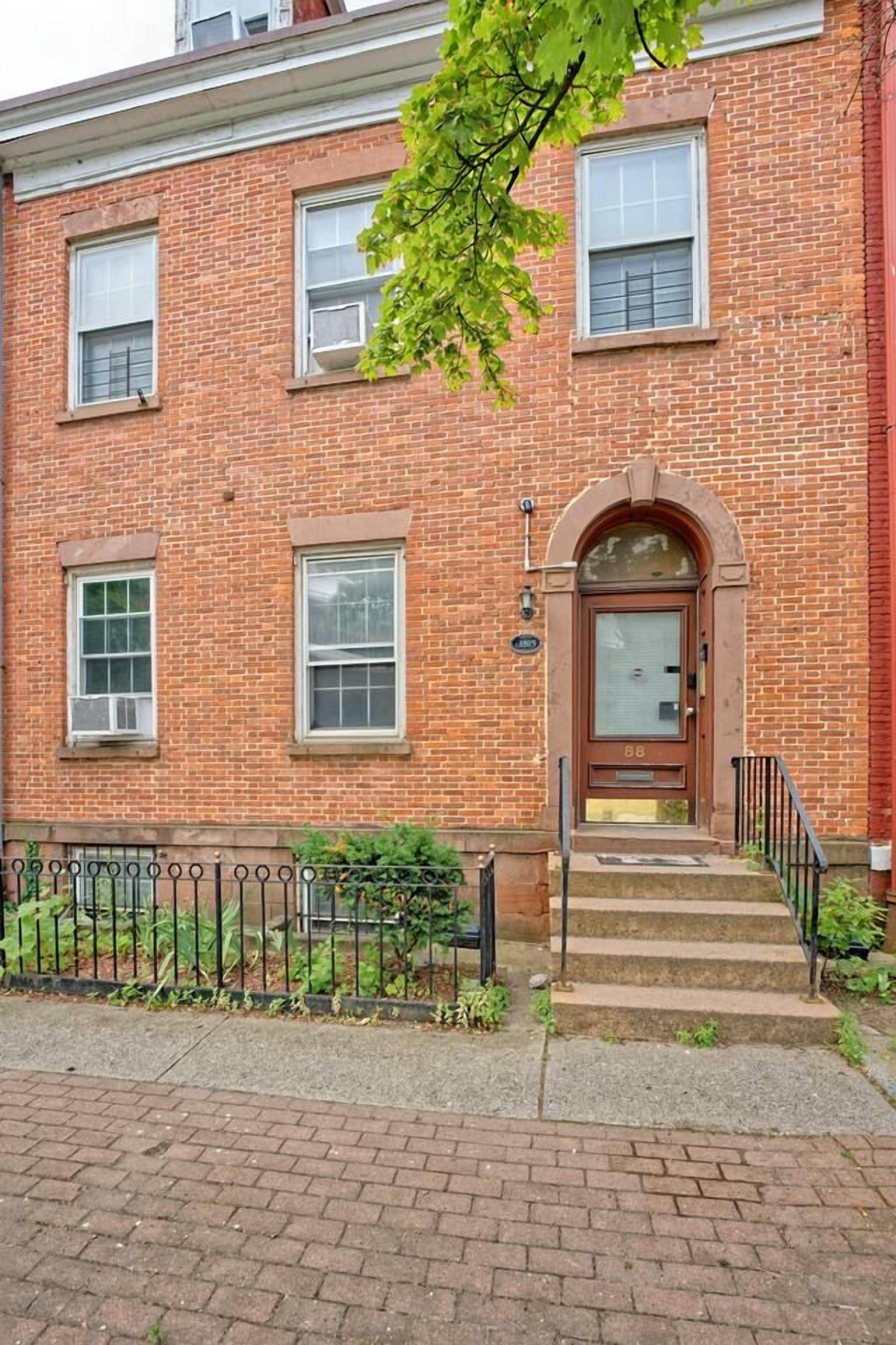 A red brick building with a central entrance, a small porch, and a metal fence.