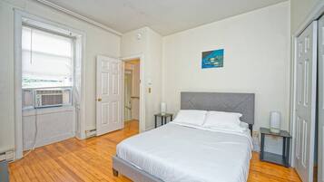 A bedroom with a bed, bedside tables, a window with blinds, a radiator, and a door leading to another room.