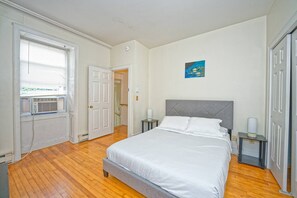 Una camera da letto con un letto, comodini, una finestra con persiane, un termosifone e una porta che dà su un'altra stanza.