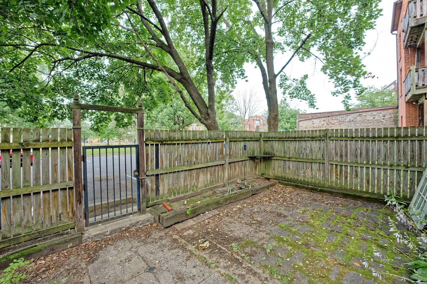 A fenced outdoor area with a paved surface, trees, and a building in the background.