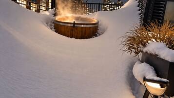 A snow-covered outdoor area with a wooden hot tub and a metal bowl, surrounded by buildings.
