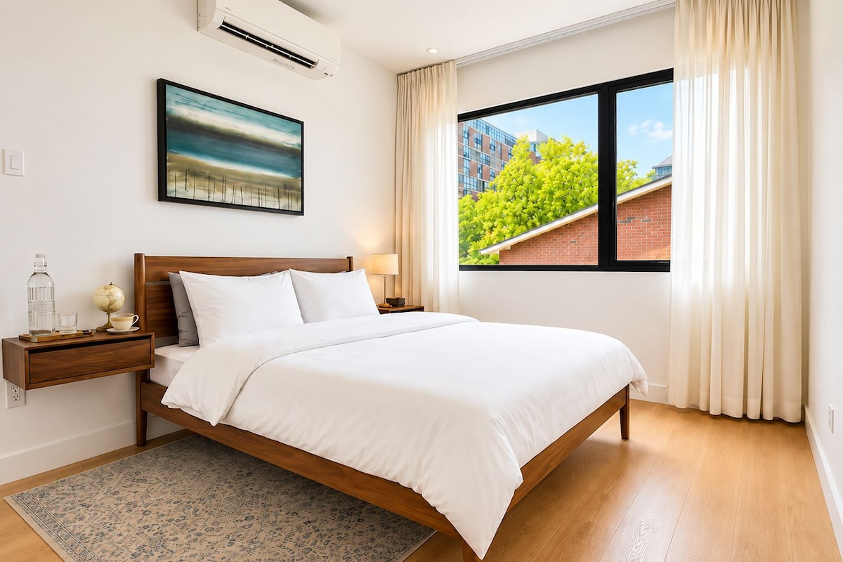 A hotel room with a large bed, a nightstand, a globe, a lamp, and a window with a view of buildings and trees.