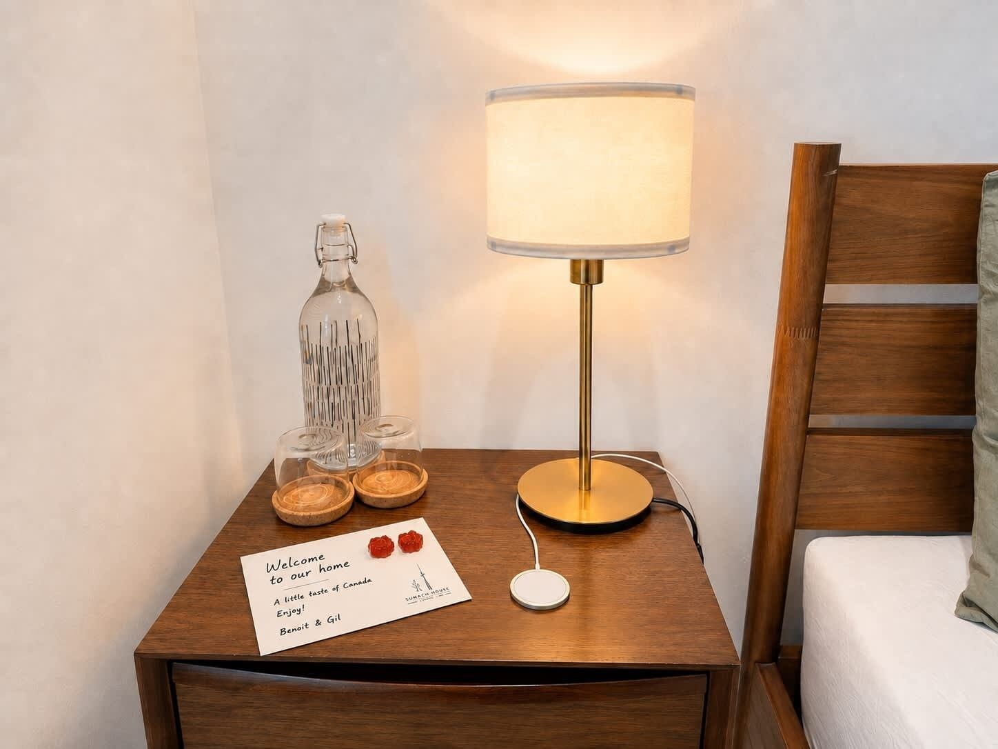 A wooden bedside table with a lamp, a glass bottle, and a welcome card.