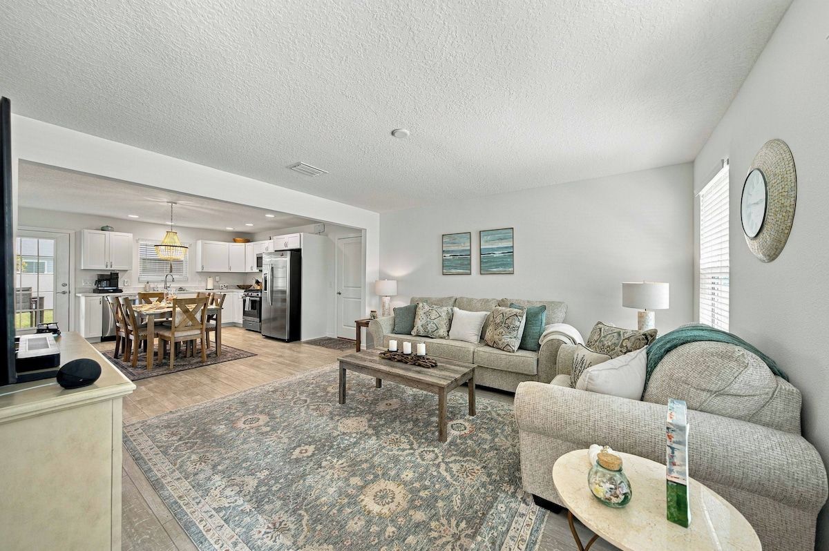 A spacious living area with a sofa, coffee table, and a rug. A kitchen is visible in the background with white cabinets and a dining area.