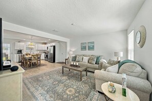 A spacious living area with a sofa, coffee table, and a rug. A kitchen is visible in the background with white cabinets and a dining area. - Brand-New 3BR · Golf Cart · Fenced Yard · Eastport (Sumterville)