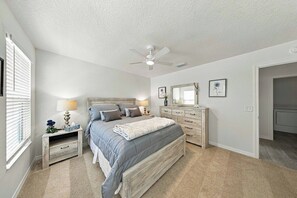 Une chambre à coucher comprenant un lit, des tables de chevet, une commode, un ventilateur de plafond et une fenêtre avec des stores.