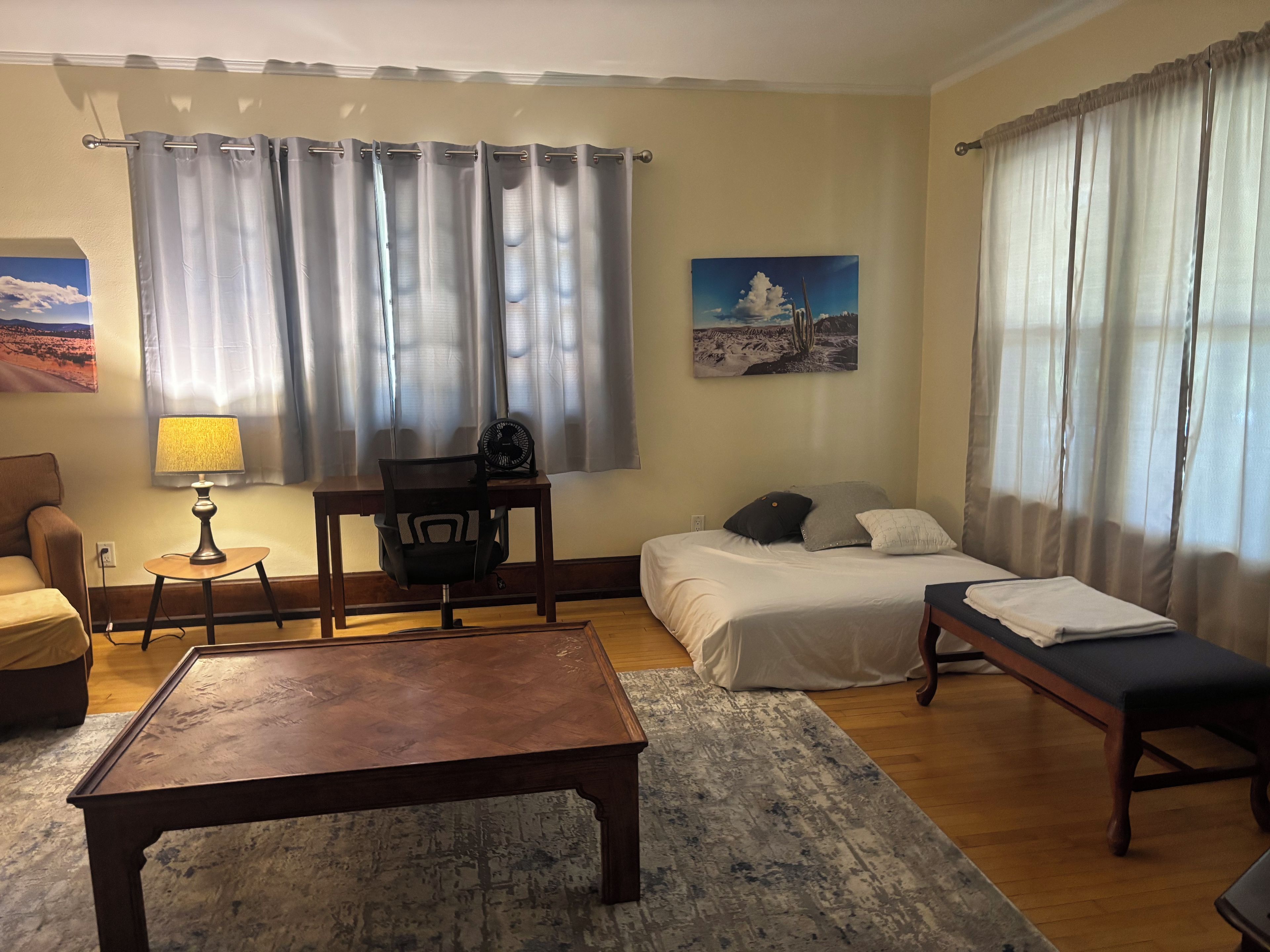 A hotel room with a bed, a desk with a chair, a small table, a bench, and a standing fan.