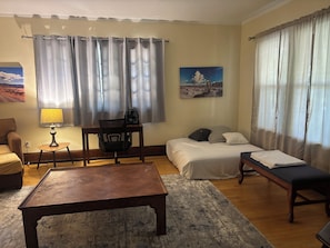 Una habitación de hotel con una cama, un escritorio con una silla, una mesita pequeña, un banco y un ventilador de pie.