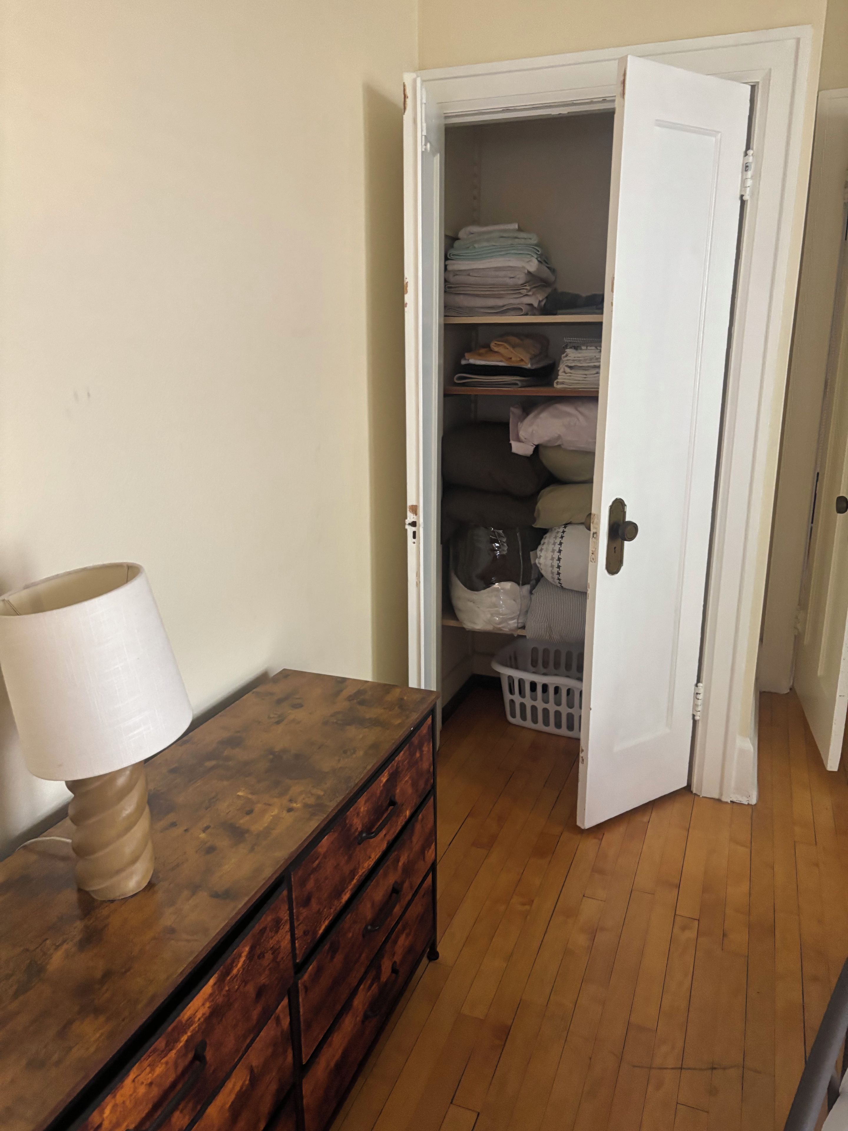 A room with a wooden dresser, a lamp, and a white door leading to a closet with shelves and a laundry basket.