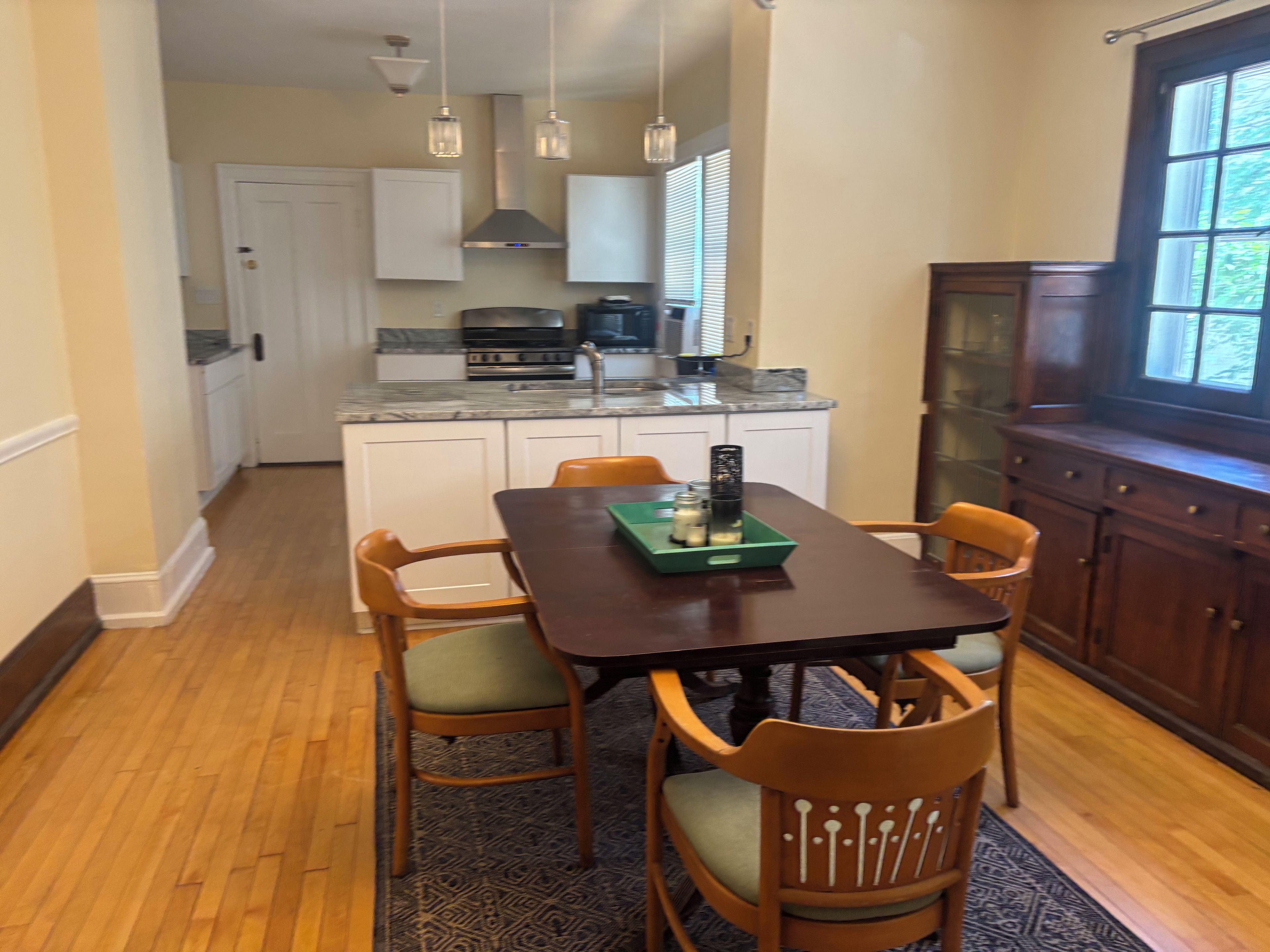 A dining area with a wooden table and chairs, a kitchen in the background, and a window with blinds.