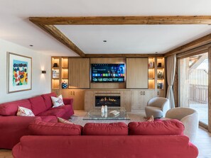A living room with a red sofa, a glass coffee table, a fireplace, wooden cabinets, and a mounted TV. - Luxury apartment, mountain view - 8 guests (Méribel)