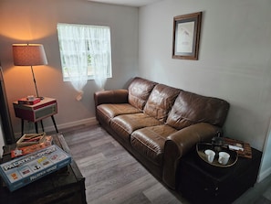 A living room with a brown leather sofa, a lamp, a framed picture on the wall, and a coffee table with a board game. - Welcome to Gator Bait Getaway! Tucked among the pines near Salt Springs. (Fort McCoy)