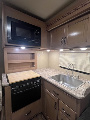 A compact kitchen with a black stove, a stainless steel sink, wooden cabinets, and a microwave. - Thor Motor Coach Bunkhouse non stationary. 
Monarch Meadows RV (Greenfield)