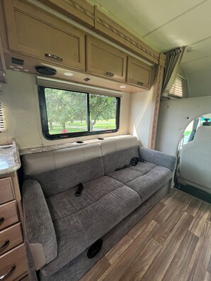 A compact RV interior with a sofa, cabinets, and a window offering a view of greenery. - Thor Motor Coach Bunkhouse non stationary. 
Monarch Meadows RV (Greenfield)