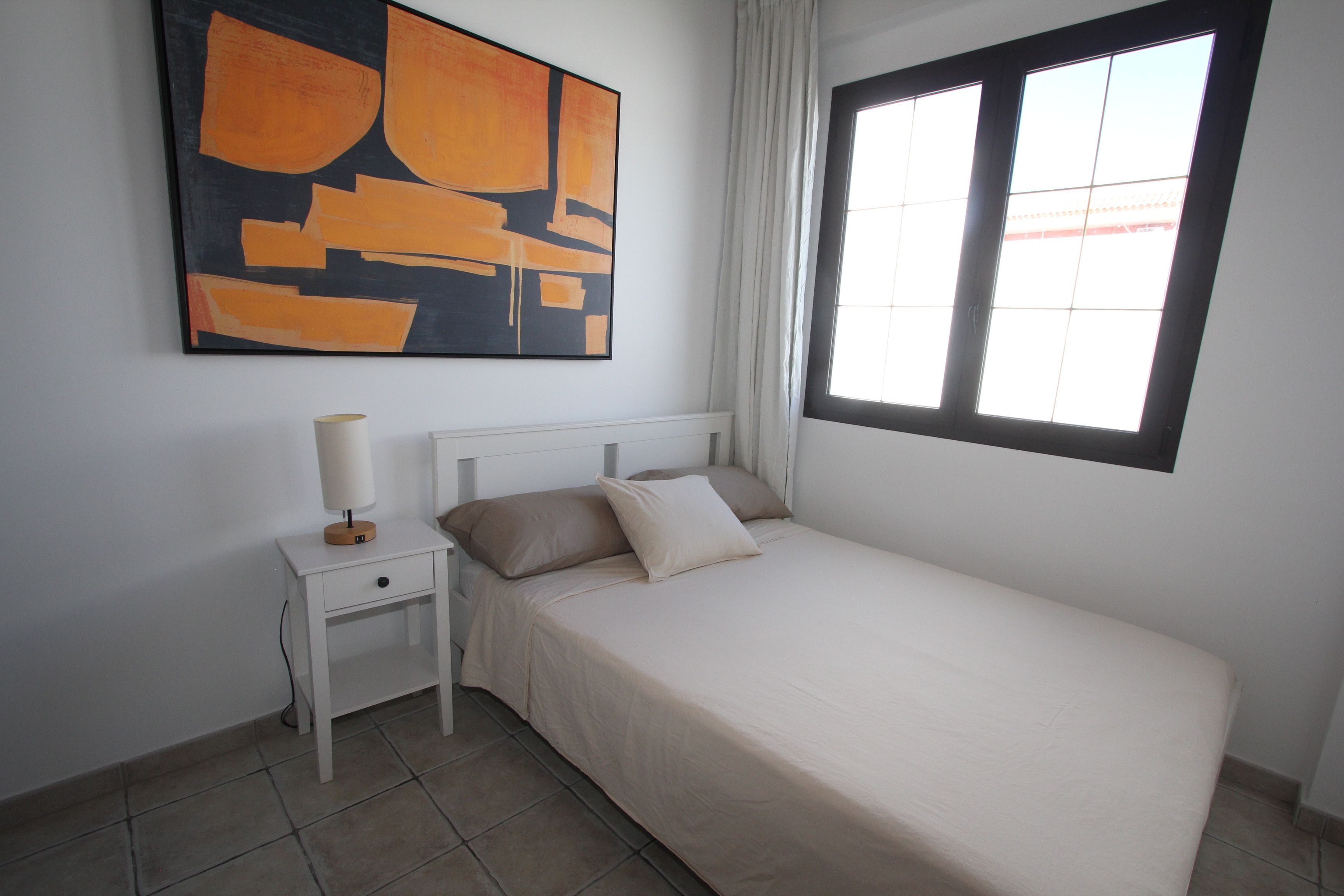 Une chambre à coucher avec un lit, une table de chevet, une lampe, une fenêtre et un tableau au mur.