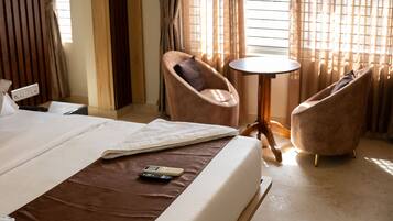 Una habitación de hotel con una cama, dos sillones, una mesita redonda y un ventanal con cortinas.