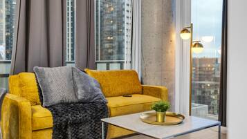 Un sofá amarillo con una manta gris, una mesita con una planta y vistas a edificios a través de amplias ventanas.