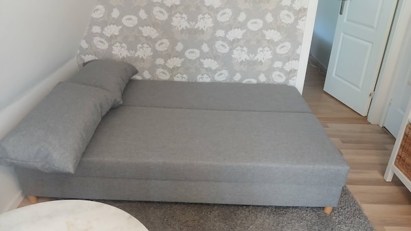 A single-bed room with a grey mattress, a grey pillow, a patterned curtain, and a wooden floor.